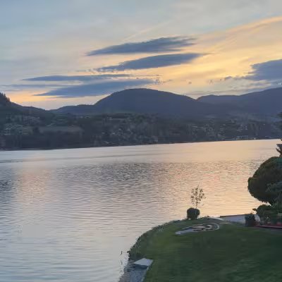 Sunset over an Okanagan lake with soft pastel sky, forested hills, and manicured shoreline