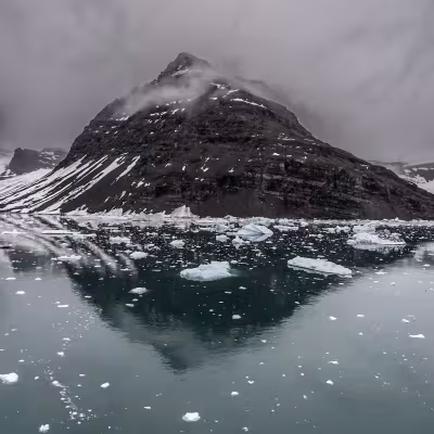 Dramatic snow-streaked mountain reflected in still, icy water, scattered with floating ice chunks under a low gray sky.