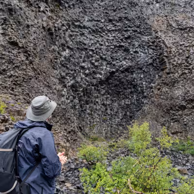 Expedition guide in rain gear and backpack standing at the base of a towering basalt rock wall, observing the geometric columns.