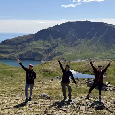Three hikers posing with arms raised on a grassy ridge above a blue bay, surrounded by steep green mountains under a bright sky.