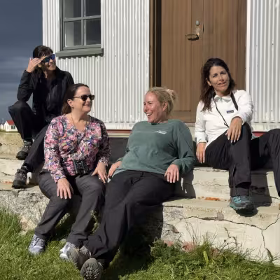 Four women sitting on the sunlit steps of a simple building, laughing and relaxing in outdoor gear with a stormy sky and yellow flowers in the background.
