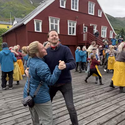Woman dancing and laughing with a local man on a wooden dock in front of a red historic building during a lively herring museum performance.