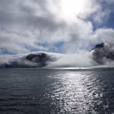 Sunlight glints off a calm Arctic bay as low clouds and fog wrap around dark, rugged mountains rising from the water.