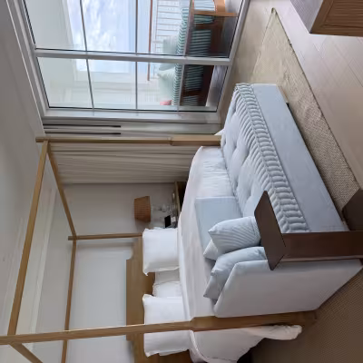 Elegant oceanfront hotel bedroom with a wooden four-poster bed, pale blue upholstered bench, and sliding doors opening to a balcony with a striped outdoor daybed and sea views