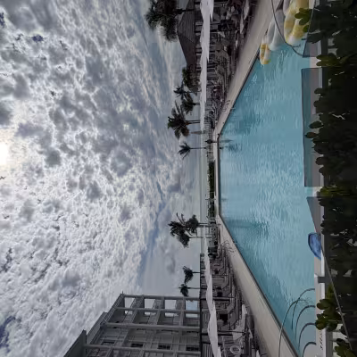 Expansive resort pool framed by lounge chairs and white umbrellas, with palm trees and the ocean in the distance under a dramatic, cloud-filled sky