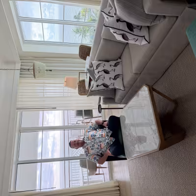 Bright, coastal-style hotel living room with large windows and balcony views of the ocean, a gray sofa with bird-print pillows, a marble coffee table, and a man seated in an armchair.