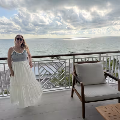 Woman in a white maxi dress and sunglasses smiling on a balcony overlooking the ocean, with a wooden lounge chair and cloudy sky above the calm water