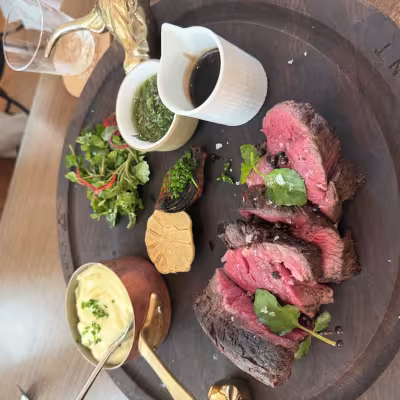 Sliced medium-rare Chateaubriand steak on a wooden board with mashed potatoes in a copper pot, chimichurri, jus, roasted garlic, and a small green salad
