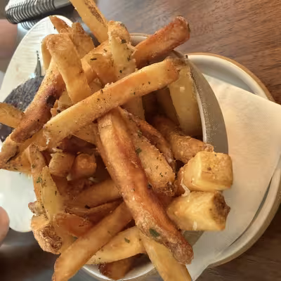 Bowl of golden, thick-cut French fries piled high on a restaurant table, with a diner in a white top holding a fry in the background