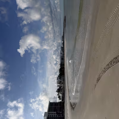 Wide sandy beach with gentle waves, tire tracks in the sand, and a line of beachfront buildings in the distance under a bright blue sky with scattered clouds
