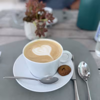 Close-up of a latte in a white cup with heart-shaped foam art, served on a saucer with a small cookie and silver cutlery on a gray placemat