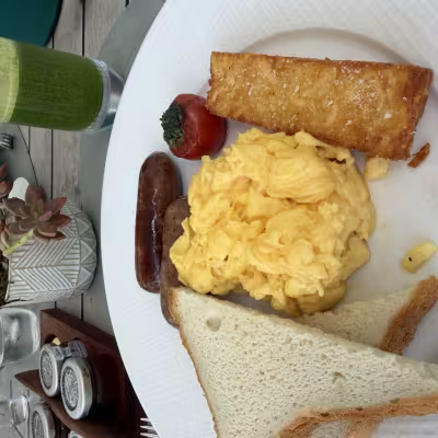 Hotel breakfast plate with scrambled eggs, sausage links, toast, and a crispy hash brown, served outdoors with a green juice and small potted succulent on the table