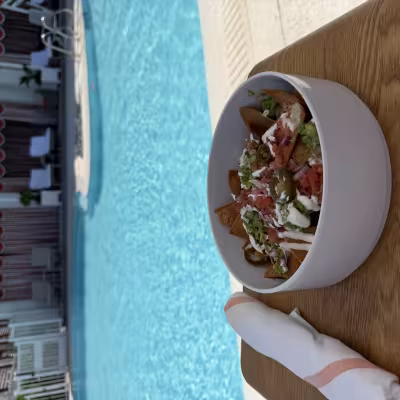 Bowl of loaded nachos topped with guacamole, jalapeños, tomatoes, onions, and crema on a wooden side table beside a bright blue resort pool and private cabanas