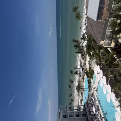 Aerial view of a luxury beachfront resort with multiple turquoise pools, rows of white umbrellas, and palm trees leading down to a calm turquoise ocean under a clear blue sky
