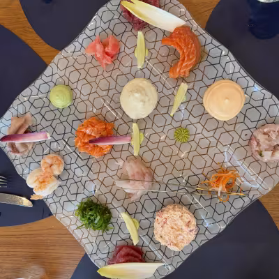 Artful seafood platter on a patterned glass plate with assorted sashimi, tartares, sauces, and garnishes arranged in a circle on a wooden restaurant table