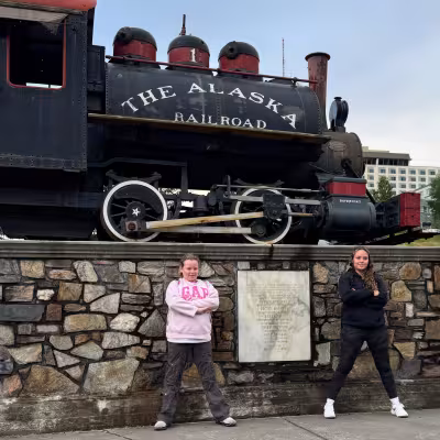 Teen girls posing by a historic Alaska Railroad steam locomotive in Anchorage before boarding the scenic train to Seward