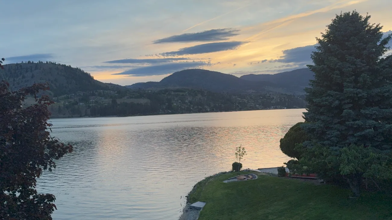 Sunset over an Okanagan lake with soft pastel sky, forested hills, and manicured shoreline