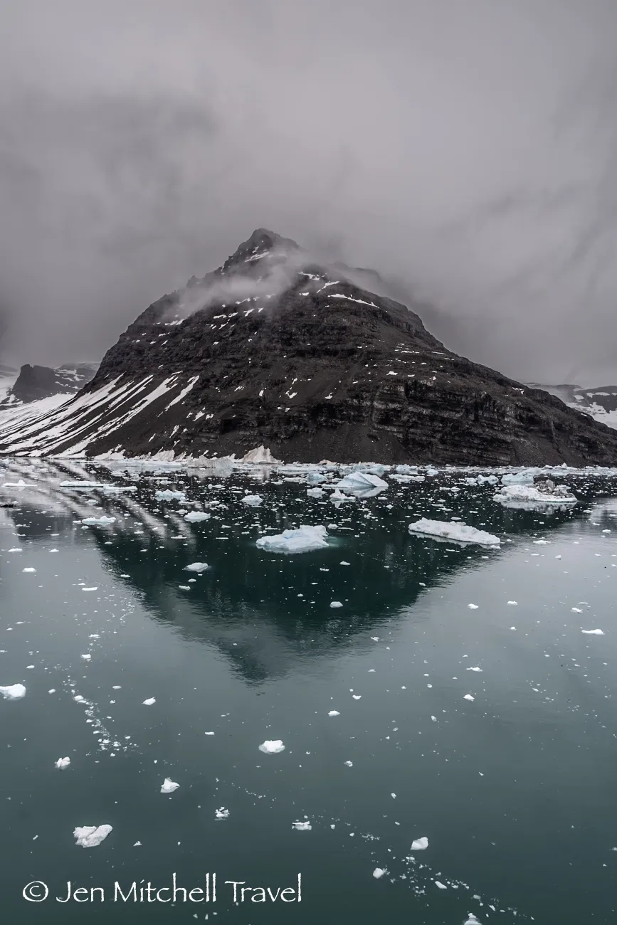 Dramatic snow-streaked mountain reflected in still, icy water, scattered with floating ice chunks under a low gray sky.