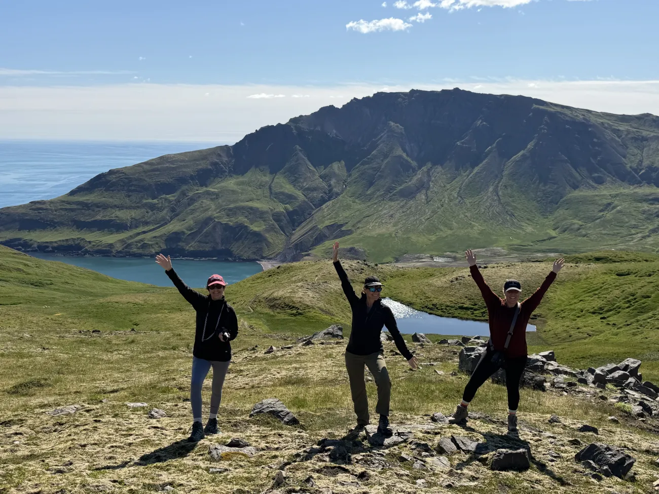 Three hikers posing with arms raised on a grassy ridge above a blue bay, surrounded by steep green mountains under a bright sky.